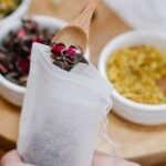 Hands filling a tea bag with dried rosebuds using a wooden spoon, set against bowls of dried herbs.