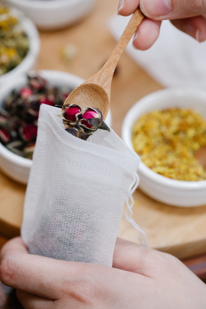 Hands filling a tea bag with dried rosebuds using a wooden spoon, set against bowls of dried herbs.